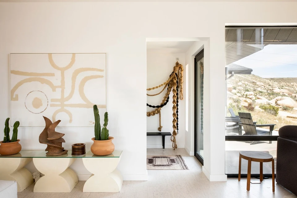 Living room interior featuring abstract art, potted cacti, and a view of a desert landscape through a large window, creating a bright, modern space.
