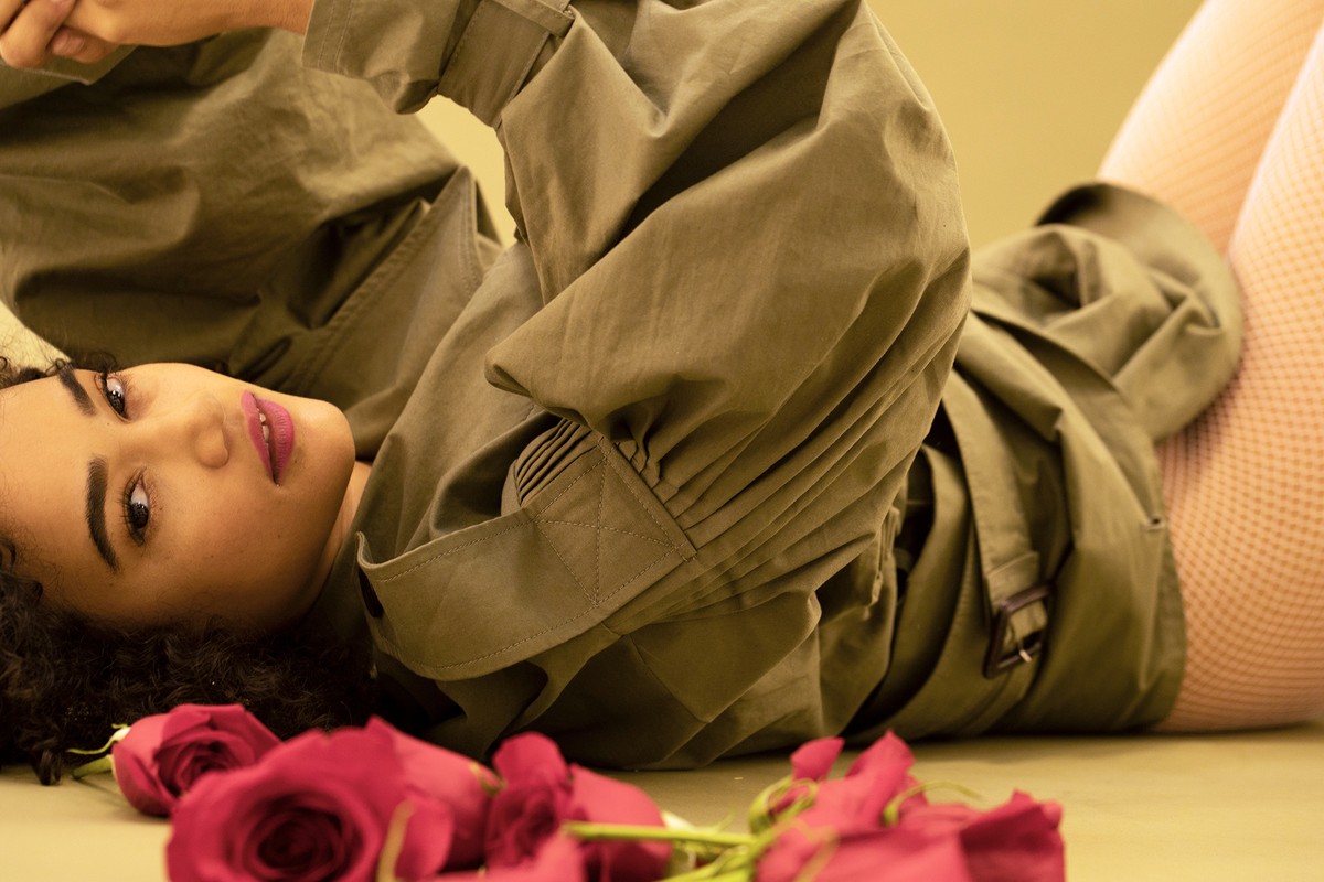 Portrait of a woman lying down in an olive-green jacket with roses in foreground and a light yellow background creating a serene mood.