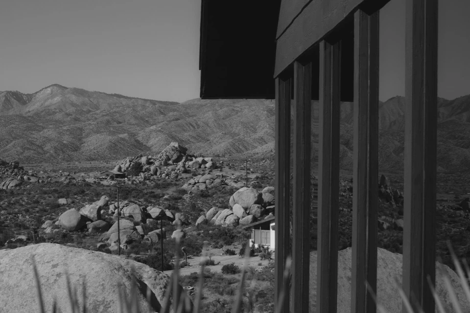Black and white landscape of rocky desert mountains, seen through the wooden beams of a building with vegetation in foreground.