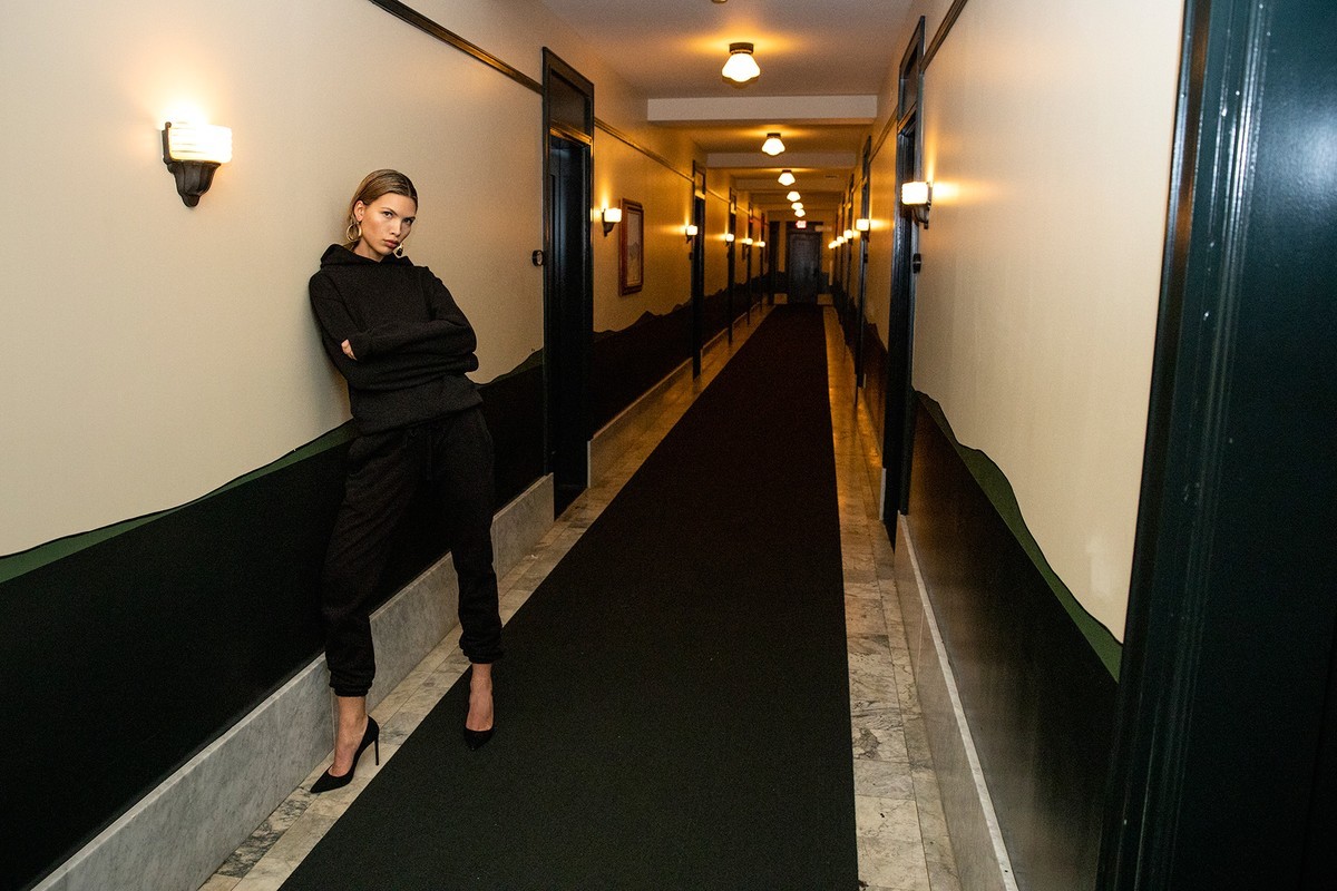 Woman in black hoodie and sweatpants posing in a hallway with black carpet and marble floors, illuminated by wall sconces.