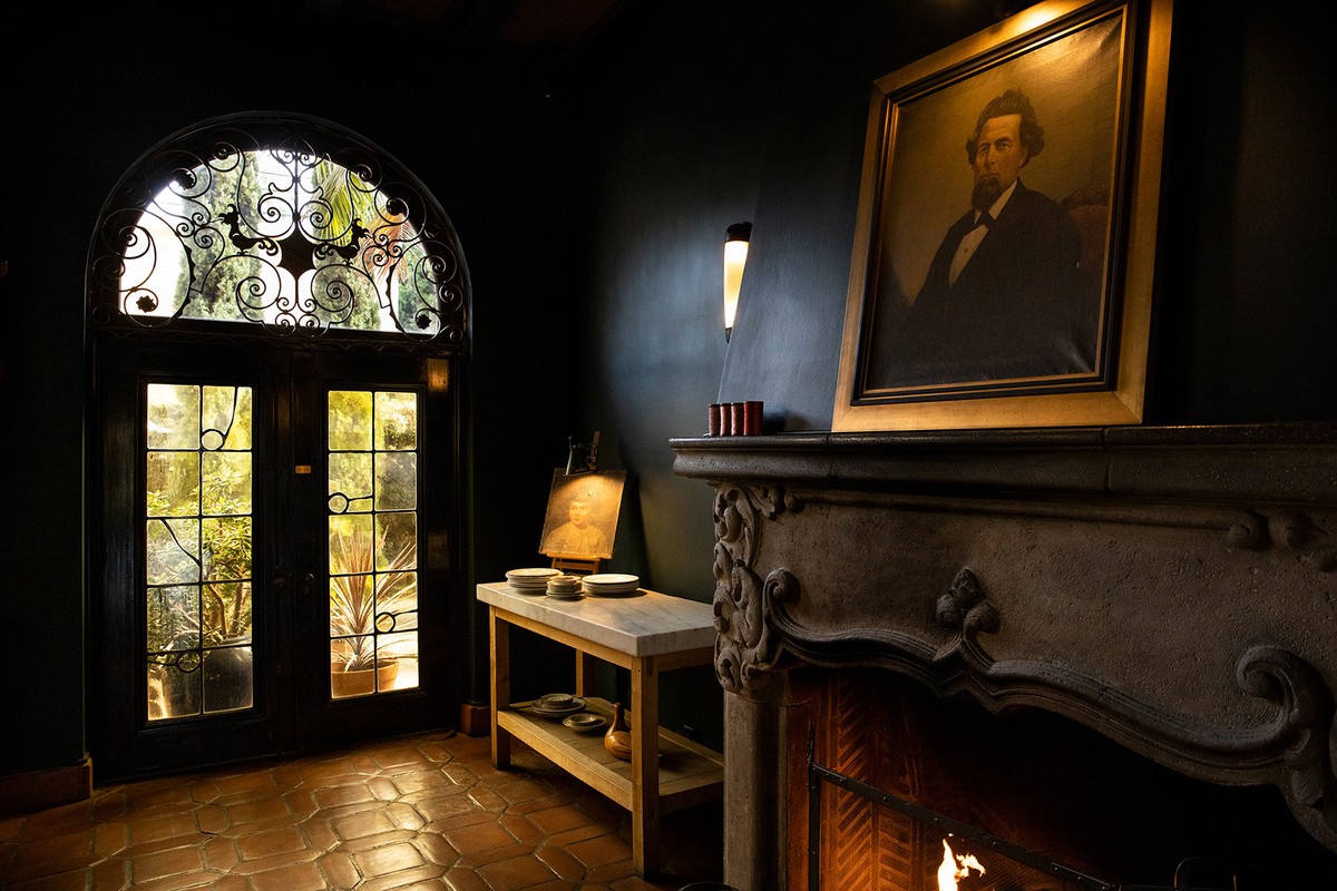 Living room interior with a portrait above a fireplace, dark walls, and a door leading to a garden with outdoor light.