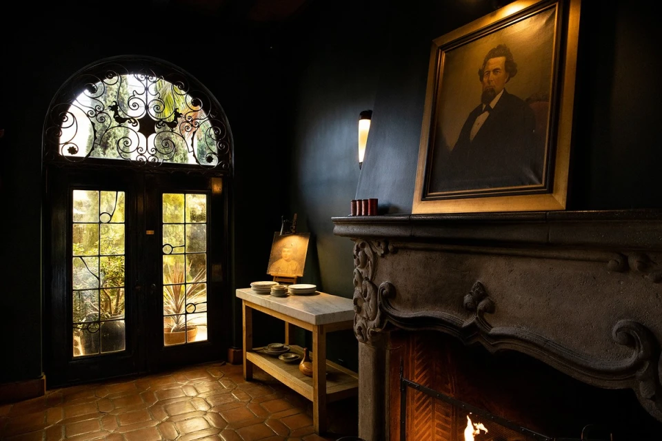 Living room interior with a portrait above a fireplace, dark walls, and a door leading to a garden with outdoor light.