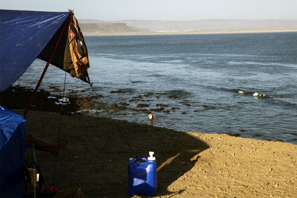 Ocean scene with a blue tarp structure on a sandy shore, calm waters, and distant mountains under a hazy sky, capturing a serene, remote ambiance.