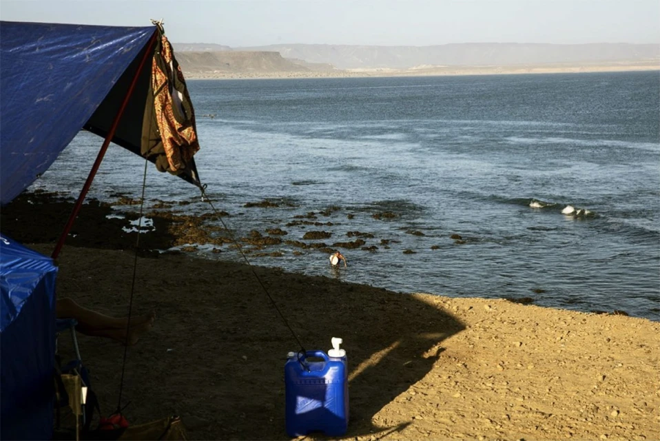 Ocean scene with a blue tarp structure on a sandy shore, calm waters, and distant mountains under a hazy sky, capturing a serene, remote ambiance.