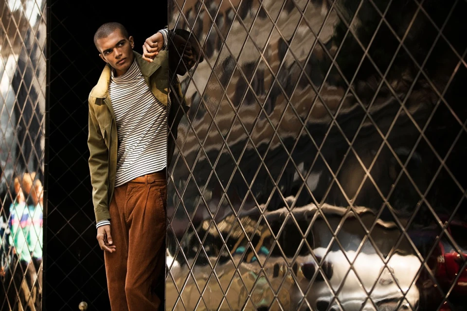 Man in striped turtleneck and corduroy pants leaning against a diamond-patterned glass wall, looking away from the camera.