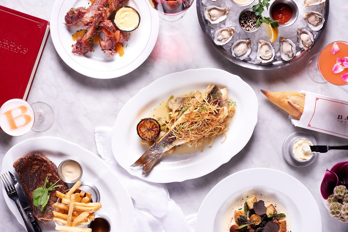 Overhead shot displays an array of dishes: grilled fish, steak with fries, oysters, baguette, and sauces on a marble table.