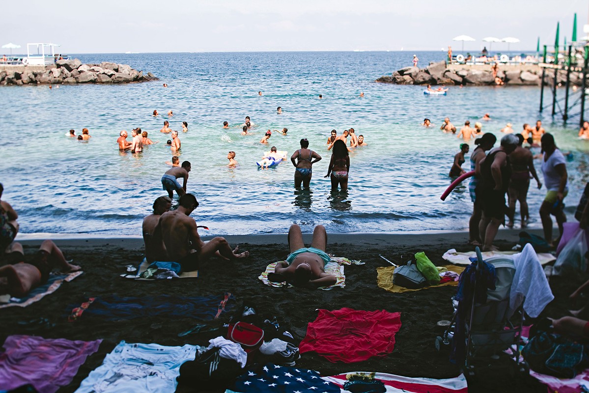 Many people swim and relax on a black sand beach with ocean views, including seated adults, sunbathers, and beach towels.