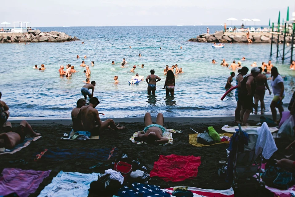Many people swim and relax on a black sand beach with ocean views, including seated adults, sunbathers, and beach towels.