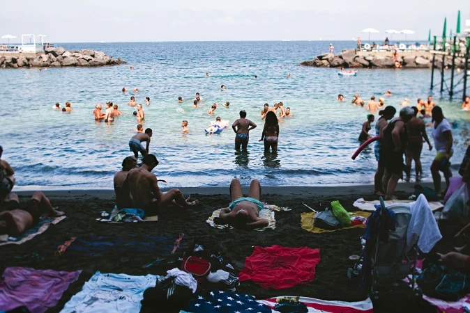 Many people swim and relax on a black sand beach with ocean views, including seated adults, sunbathers, and beach towels.
