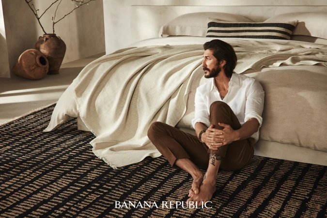 Man with beard sitting on a patterned rug against a neutral bedroom set, wearing a white shirt and brown trousers, photographed by Mike Carreiro.