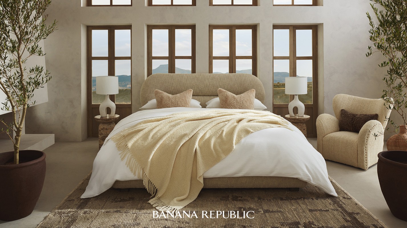 Bedroom interior with a beige bed, throw blanket, pillows, side tables with lamps, textured armchair, olive trees in planters, and multiple windows.