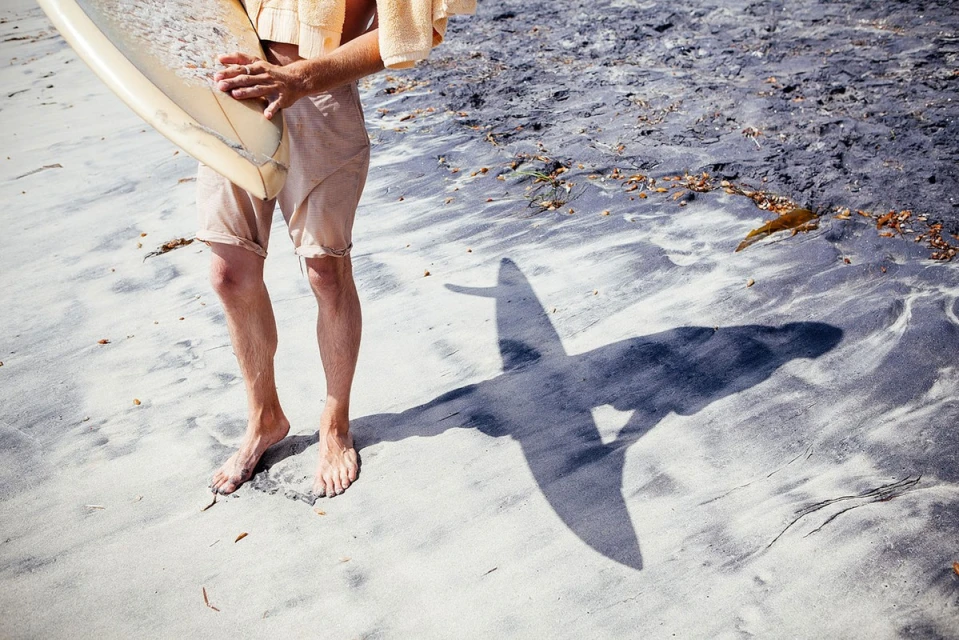 A man stands barefoot in the sand, holding a surfboard with a towel slung around his shoulders. His shadow is cast on the beach.