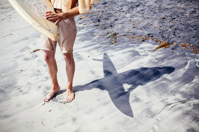 A man stands barefoot in the sand, holding a surfboard with a towel slung around his shoulders. His shadow is cast on the beach.