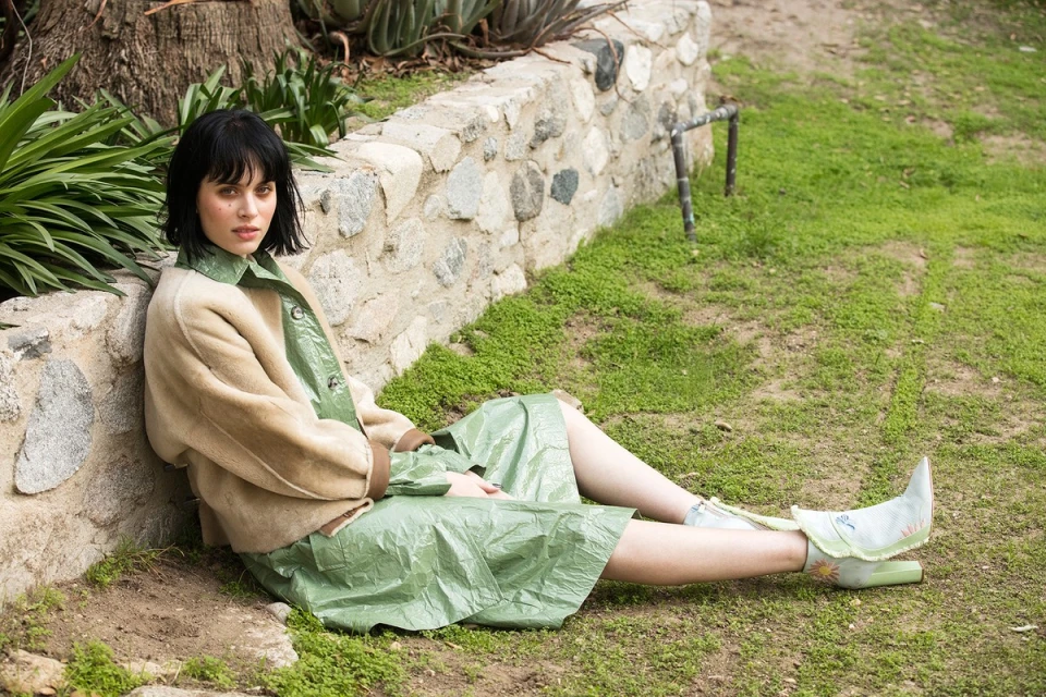 A stylish adult woman with dark hair poses in an outdoor setting, wearing a light green dress and a tan-colored jacket, sitting on the grass.