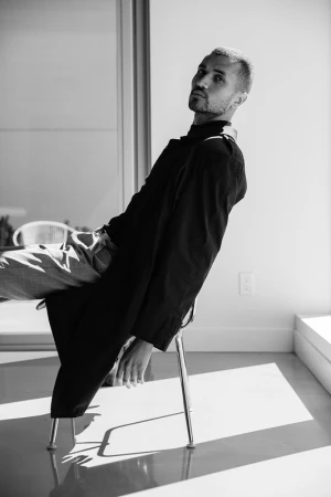 Black and white portrait of a man in a dark coat, seated on a chair by a bright window, creating contrast between light and shadow.