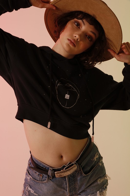A young woman wearing a black sweatshirt, distressed denim shorts, and a tan hat poses in a studio with soft, diffused lighting.