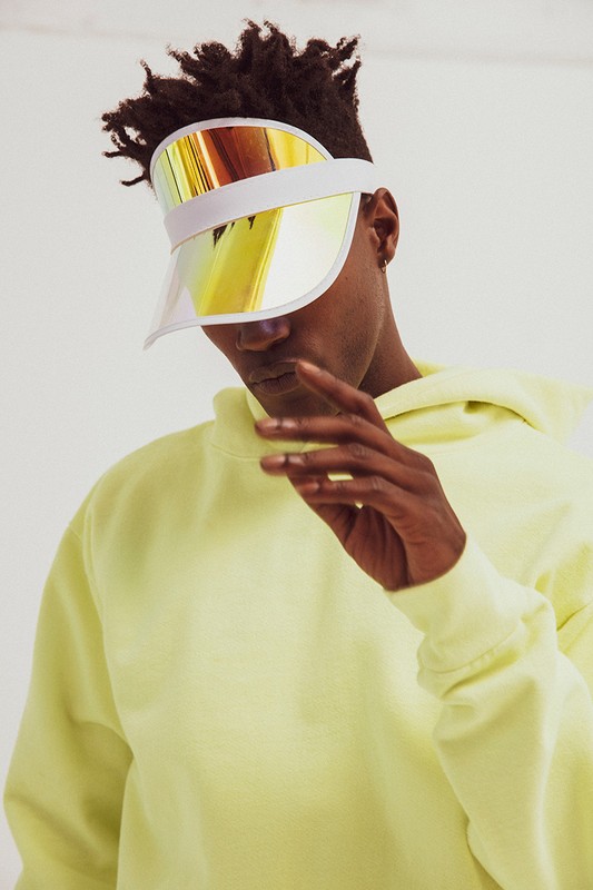 Person wearing a bright-yellow hooded sweatshirt and a reflective visor, against a white background, in a studio shot.