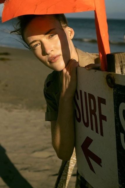 Portrait of a woman with light skin at the beach, next to a weather-beaten surfboard sign and orange buoy on a sunny day.