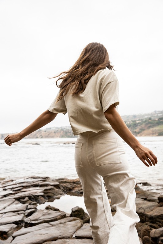 A woman stands on rocks by the ocean with outstretched arms, wearing a beige crop top and white denim pants on an overcast day.