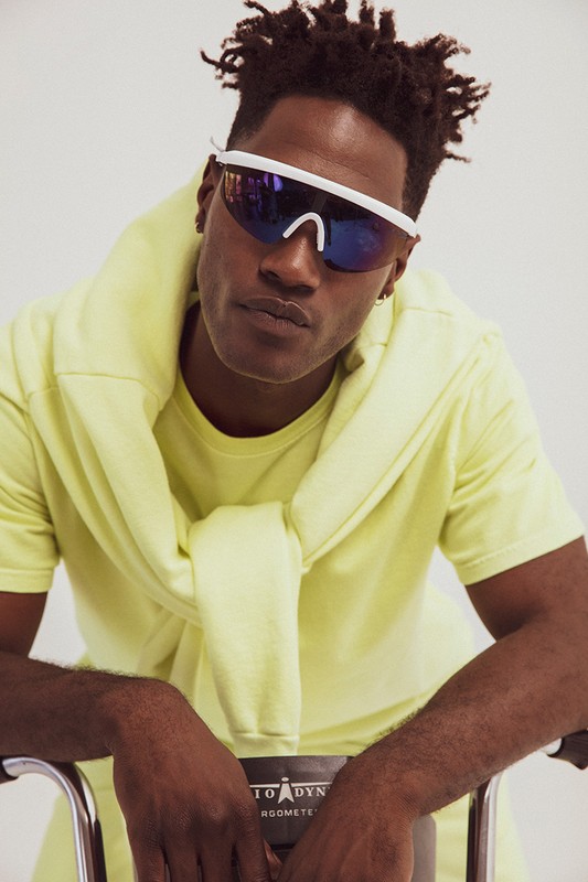 Portrait of a dark-skinned male model in neon yellow sportswear and white wraparound sunglasses against a bright, neutral backdrop.