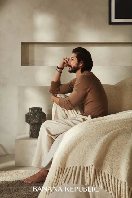 A bearded man sits on a bed in a beige room, wearing a brown sweater and cream pants in an indoor fashion setting.