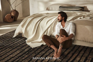 Man with beard sitting on a patterned rug against a neutral bedroom set, wearing a white shirt and brown trousers, photographed by Mike Carreiro.