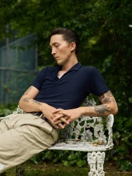 A tattooed man with slicked hair wearing a dark polo and khaki trousers seated outdoors on an ornate white garden bench.