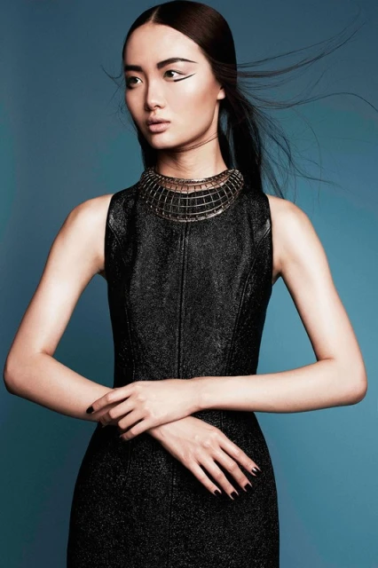 Elegant Asian woman in a black dress with a striking necklace, posing against a serene teal backdrop in a fashion-forward portrait.