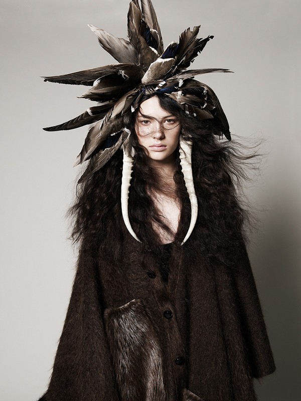 Portrait of a woman with elaborate feathered headdress and faux tusks, wearing a dark brown coat, posing in a neutral studio setting.