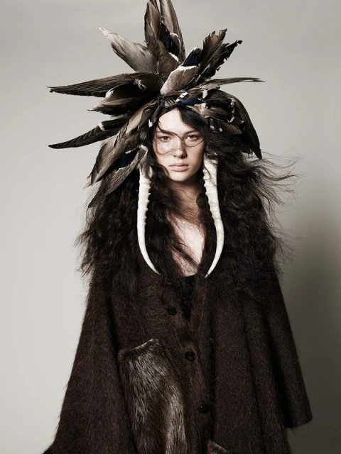 Portrait of a woman with elaborate feathered headdress and faux tusks, wearing a dark brown coat, posing in a neutral studio setting.