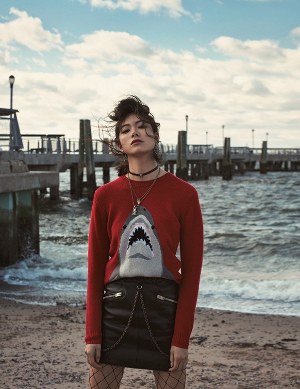A young woman stands on a beach near a pier in a red shark sweater and black skirt, exuding a casual yet fashionable vibe on a cloudy day.