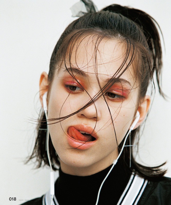 Close-up portrait of a young woman with wet hair and colorful eye makeup, wearing headphones and sticking her tongue out against a white background.