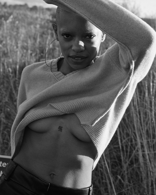Portrait of a Black female with shaved head in v-neck sweater, lifting the garment to show upper body and small tattoo. Outdoors setting.