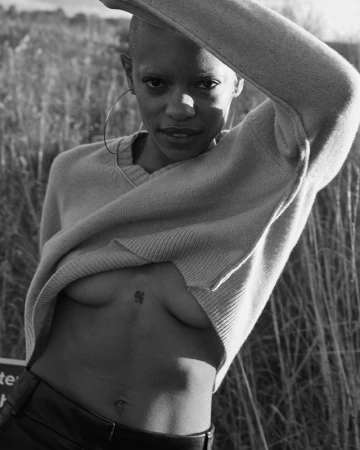 Portrait of a Black female with shaved head in v-neck sweater, lifting the garment to show upper body and small tattoo. Outdoors setting.