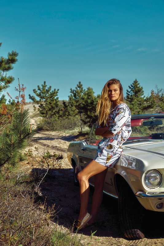 Model leaning against a vintage convertible car outdoors in a landscape with sparse trees under a blue sky.