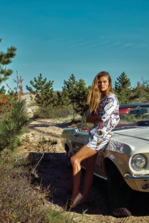 Model leaning against a vintage convertible car outdoors in a landscape with sparse trees under a blue sky.