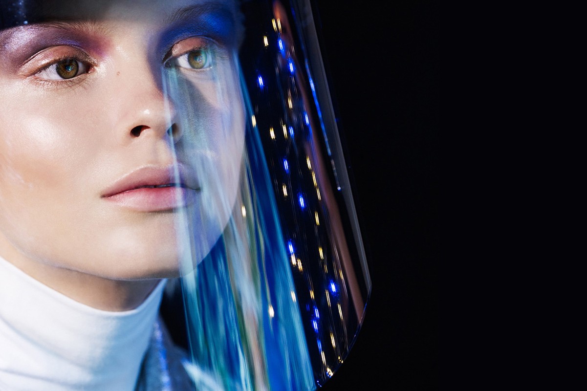 Close-up studio portrait of a woman with creative makeup and futuristic visor reflecting blue lights against a dark background.