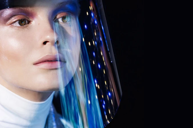 Close-up studio portrait of a woman with creative makeup and futuristic visor reflecting blue lights against a dark background.