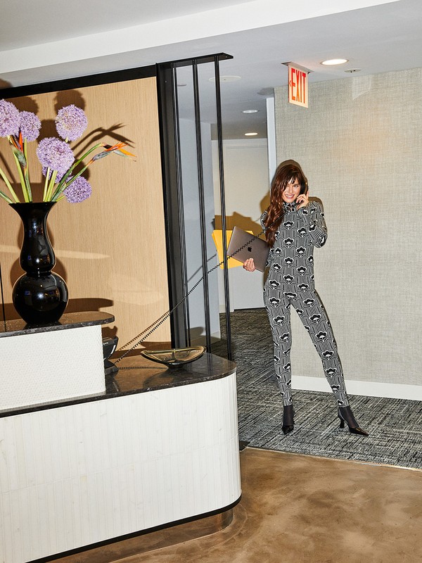 An adult woman in a patterned jumpsuit stands in a building hallway holding a laptop, with flowers and a front desk in the background.