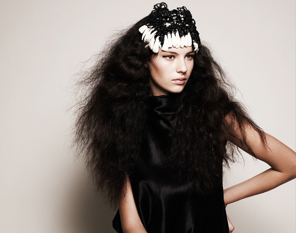 Portrait of a young woman with long, dark, voluminous hair wearing a unique black-and-white headpiece and a black top in a studio shot.