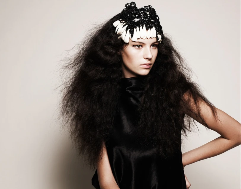 Portrait of a young woman with long, dark, voluminous hair wearing a unique black-and-white headpiece and a black top in a studio shot.