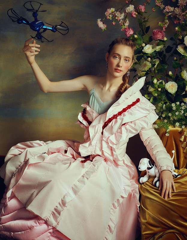Woman in a ruffled pink gown holds a drone in a fine art style, floral background, and robotic dog prop, in a studio setting.