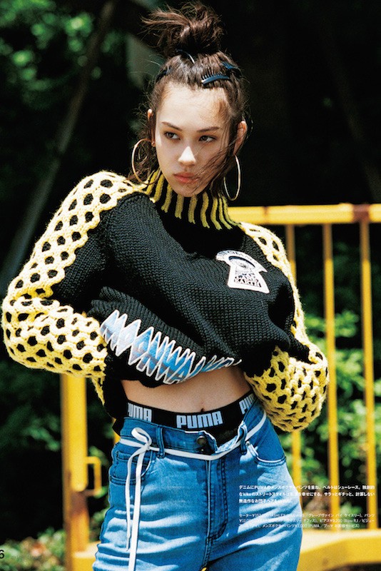 Fashion editorial portrait of a young adult woman in a black and yellow sweater, denim jeans, and hoop earrings outdoors.