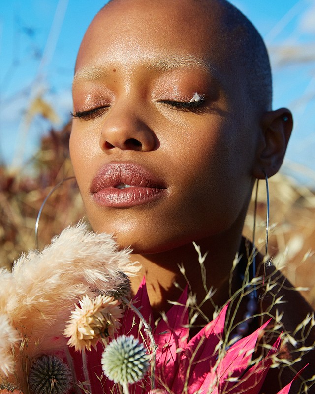 Close-up portrait of a bald woman with closed eyes, holding colorful flowers against an outdoor background