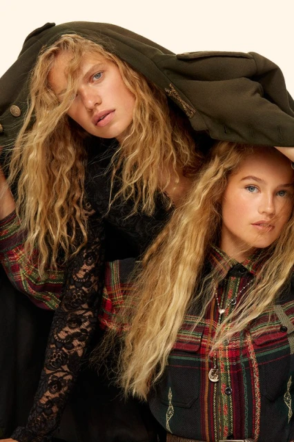 Two women pose closely, one in olive coat, one in plaid shirt, sharing a natural blonde hairstyle and similar facial features in a studio setting.