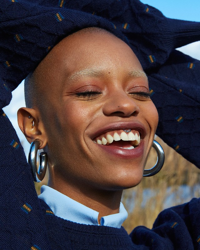 Smiling Black woman with shaved head wearing silver hoop earrings and a blue patterned sweater posing outdoors against a natural background.