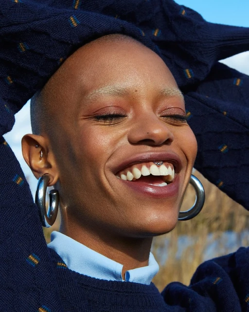 Smiling Black woman with shaved head wearing silver hoop earrings and a blue patterned sweater posing outdoors against a natural background.
