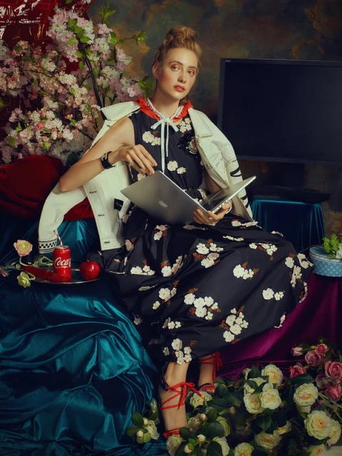 Young woman in floral dress sits with laptop in opulent setting amidst roses, coca cola can, and laptop in artistic, indoor setting.