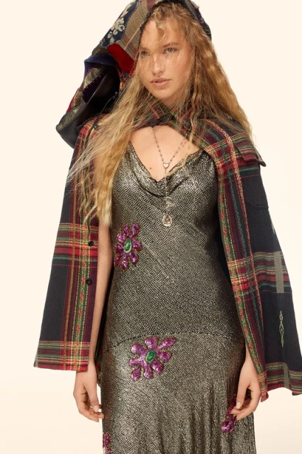Fashion shot of a blond woman in a silver dress with floral embellishments, wearing a patchwork cape, against a neutral backdrop.