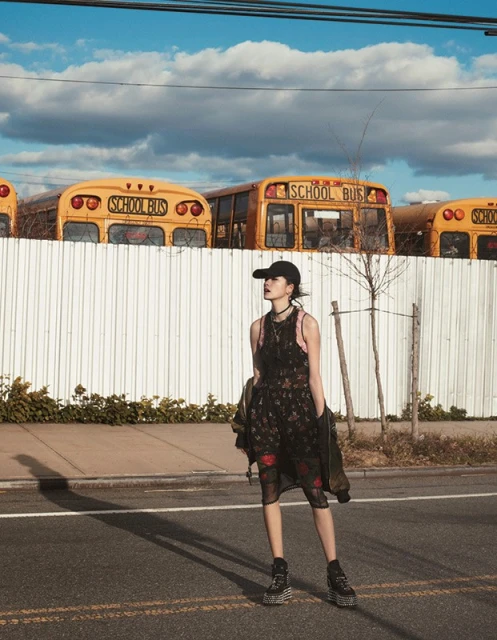 A woman in a floral dress and baseball cap stands on an asphalt road with parked yellow school buses in the background under a blue sky.